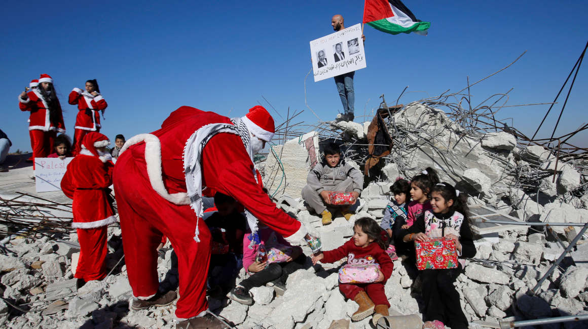 Papá Noel entrega regalos a niños sentados sobre los restos de una casa demolida en Palestina.